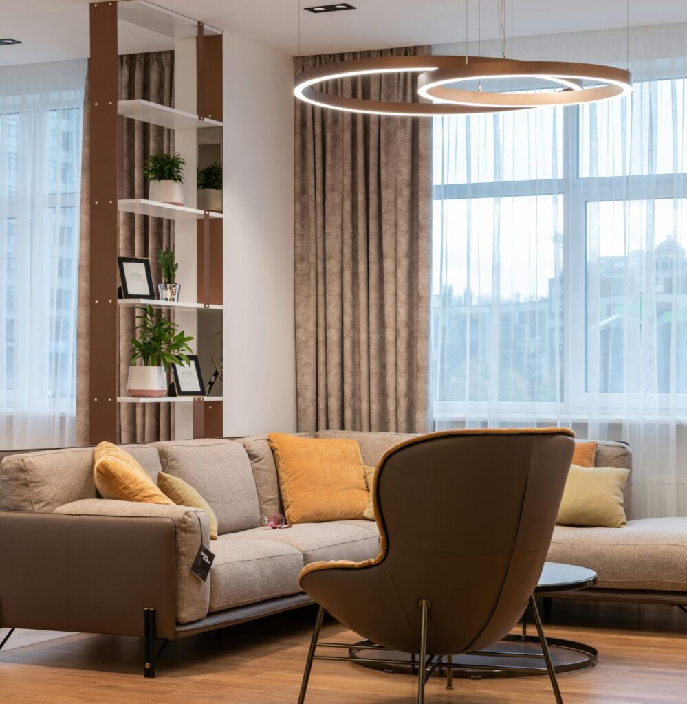 Home Modern living room interior with soft couch and pillows near armchair and round table near window with curtains and shelves with plants near lamp